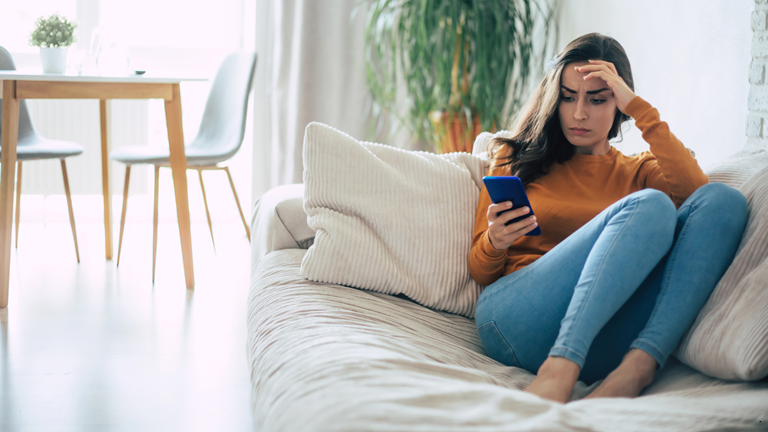 Illustration einer nachdenklichen Frau, die seitlich auf einem Sofa sitzt und auf ihr Smartphone schaut, weil er sich nicht meldet und Unsicherheit entsteht.